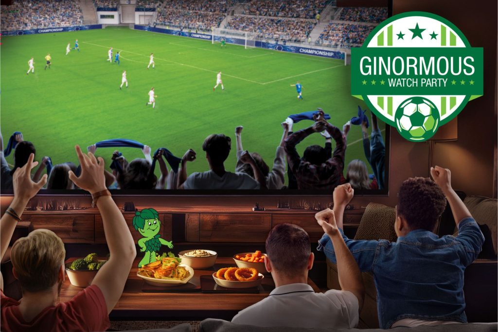 A group of friends cheers excitedly in front of a large TV displaying a soccer match, with snacks on the table and a colorful cartoon character beside them, creating a festive watch party atmosphere, featured in the Giant Watch Party Sweepstakes