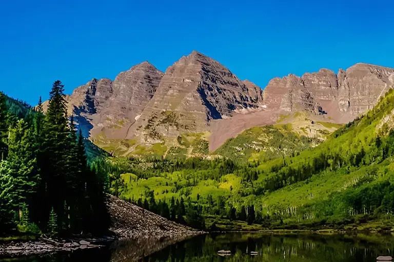 Lush green valleys surround towering mountains under a clear blue sky, reflecting beautifully in a tranquil lake, creating a serene and picturesque landscape, featured in the San Pellegrino Aspen Food & Wine Sweepstakes