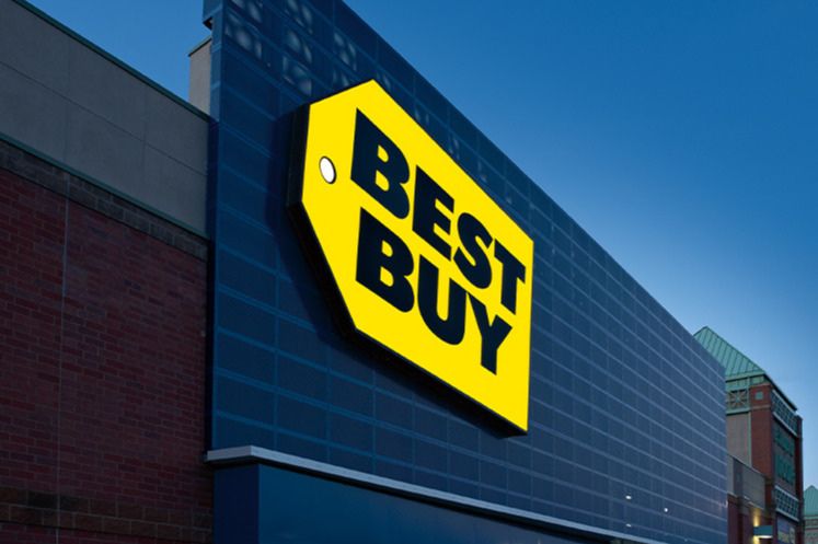 A brightly lit storefront features a large yellow and black sign against a darkening blue sky, indicating the presence of a major electronics retailer, featured in the Best Buy $1,000 Sweepstakes