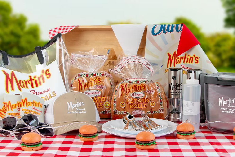 A picnic setting features a checkered tablecloth with mini burgers and various Martin's potato rolls, accompanied by sunglasses, a cap, and grilling tools against a blurred outdoor background, featured in the Martins Summer Fun Sweepstakes