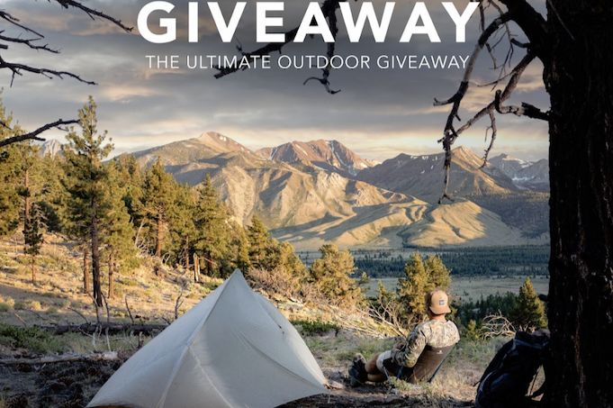 A person sits on a hillside, gazing at a stunning mountain landscape, with a tent pitched nearby, surrounded by trees in a serene outdoor setting, featured in the melin Howl R1 Fire Pit Sweepstakes