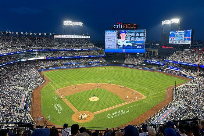 A panoramic view of a bustling baseball stadium at night, with a packed crowd watching the game under bright lights, and a large scoreboard displaying player information, featured in the GEICO Feels Good Sweepstakes