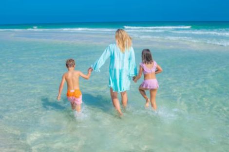 A woman and two children walk through shallow water at the beach, enjoying a sunny day as gentle waves lap around their legs. The scene conveys a sense of joy and family togetherness, featured in the Visit Pensacola Getaway Sweepstakes