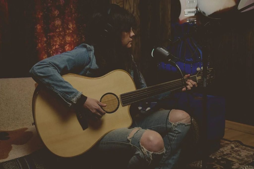 A musician plays an acoustic guitar while seated in a cozy studio, wearing headphones and focused on their performance amidst warm, colorful lighting, featured in the LR Baggs Shure Giveaway
