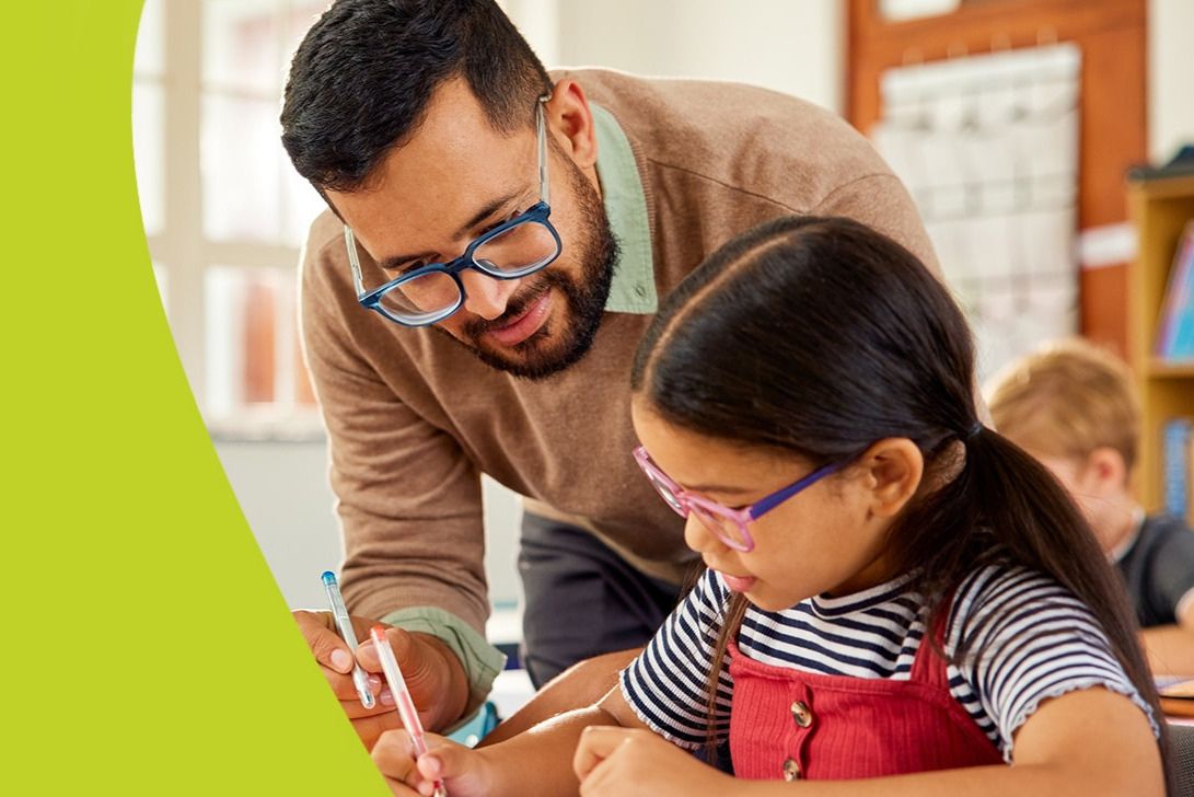 A teacher leans over to assist a young student with her writing, focusing on a worksheet. The classroom features bright natural light and other students engaged in learning, featured in the Eyemart Express $1,000 Teacher Sweepstakes