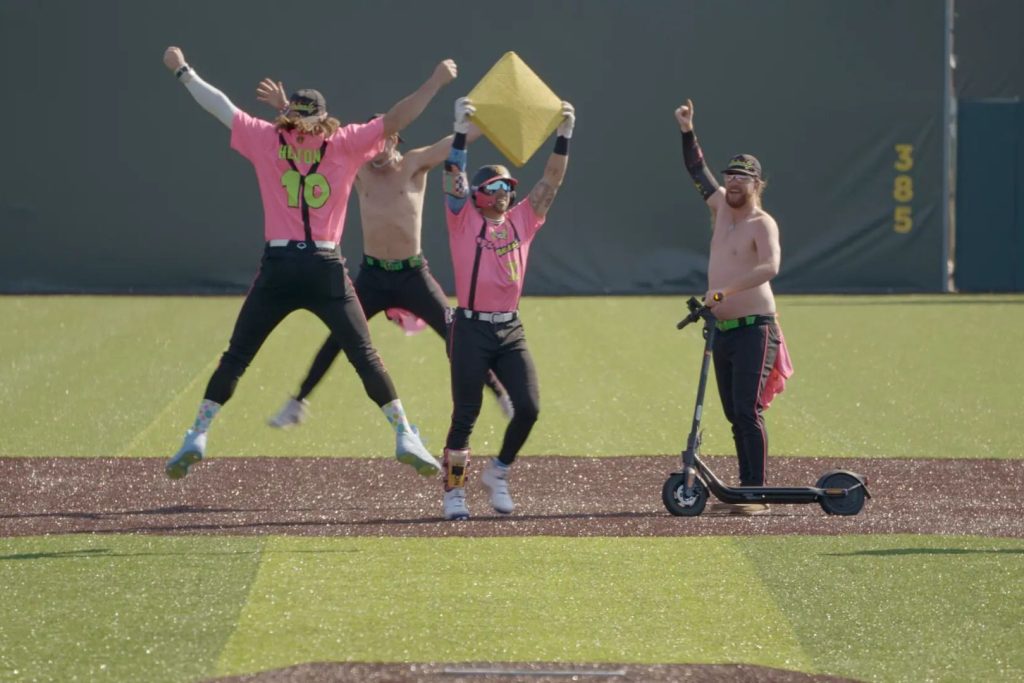 A group of four playful individuals in bright pink shirts celebrates on a baseball field, one holding a base while another rides a scooter, capturing a lively moment of enthusiasm, featured in the GEICO Banana Ball Championship Sweepstakes