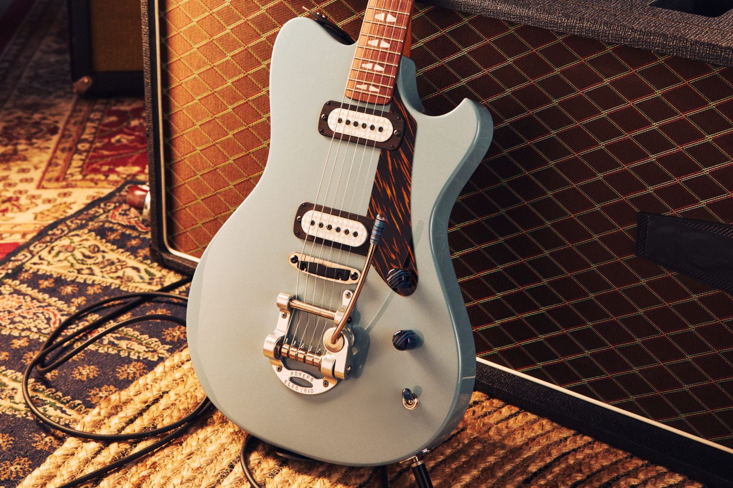 A sleek, light blue electric guitar leans against a textured amplifier, with guitar cables intertwined on a woven rug. The scene conveys a cozy music-making atmosphere, featured in the Reverb's Powers Electric Sweepstakes