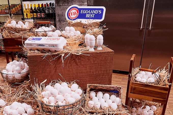 A display of baskets filled with white eggs is arranged on a wooden table, surrounded by straw, in a brightly lit grocery store setting, featured in the Eggland’s Best $5,000 Sweepstakes