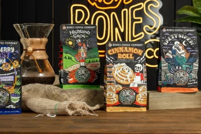 A wooden table displays three vibrant coffee bags labeled "Highland Grog," "Cinnamon Roll," and "Salty Swen," alongside a coffee maker and a burlap cloth, set against a dark background, featured in the Bones Free Coffee Sweepstakes