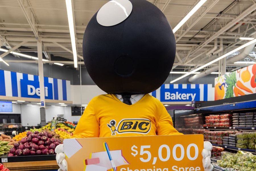 A humorous character with a large round head and a yellow shirt stands in a grocery aisle, holding a sign advertising a $5,000 shopping spree amidst colorful produce displays, featured in the BIC Teacher Shopping Spree Sweepstakes