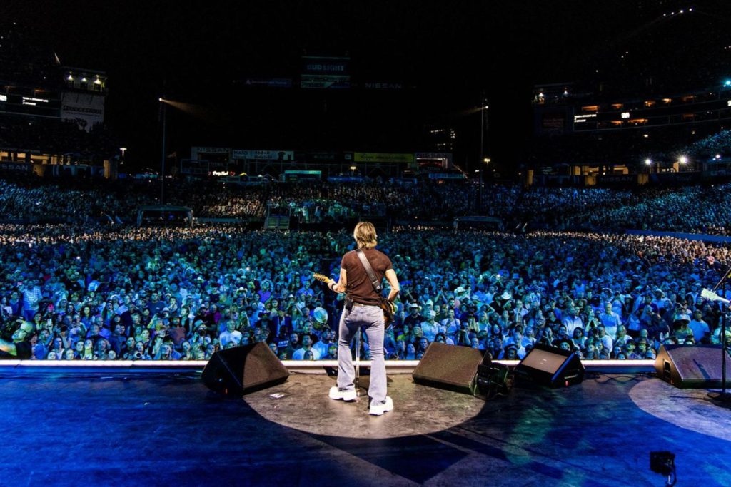 A musician stands on stage, guitar in hand, facing a large, enthusiastic crowd illuminated by colorful stage lights at a night concert, featured in the Visit Music City CMA Fest Sweepstakes