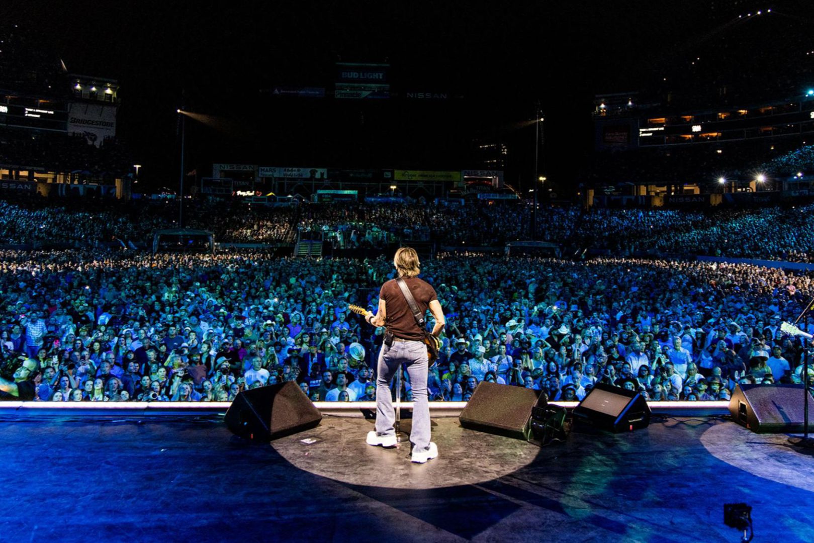 A musician stands on stage, guitar in hand, facing a large, enthusiastic crowd illuminated by colorful stage lights at a night concert, featured in the Visit Music City CMA Fest Sweepstakes