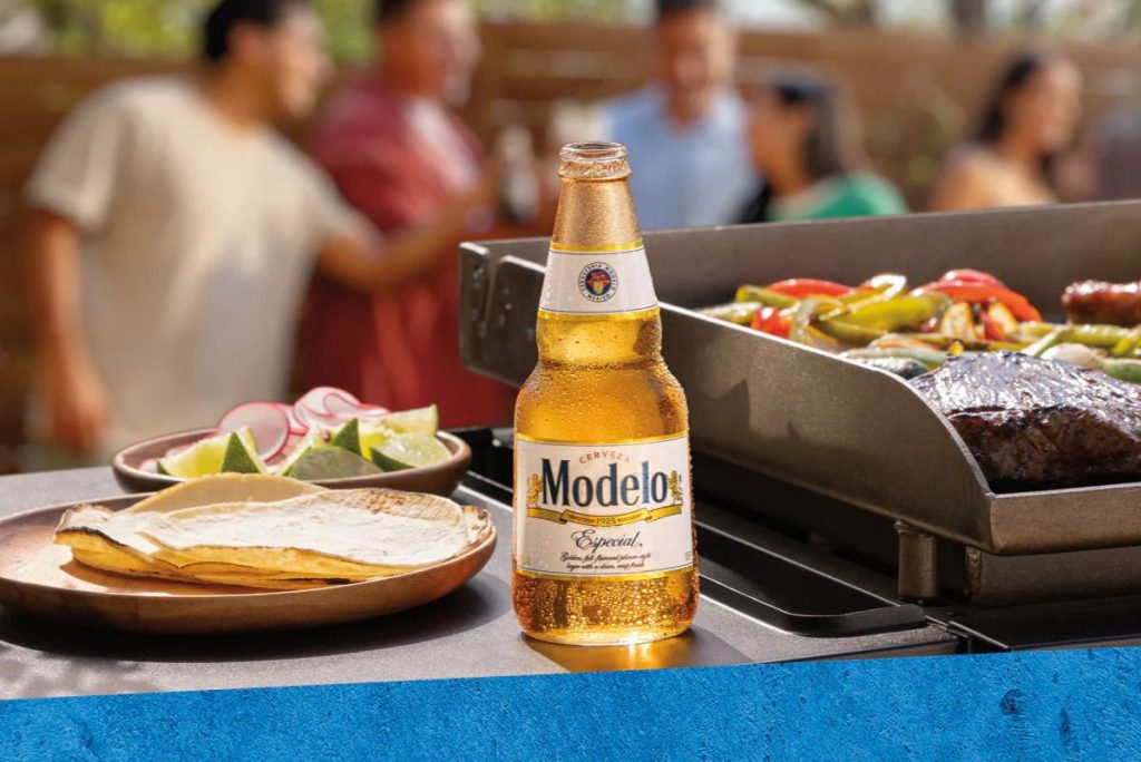 A chilled beer is prominently displayed beside a platter of tortillas and fresh lime and radish slices, while friends enjoy grilling meat and vegetables in the background, featured in the Modelo Summer 2026 Sweepstakes