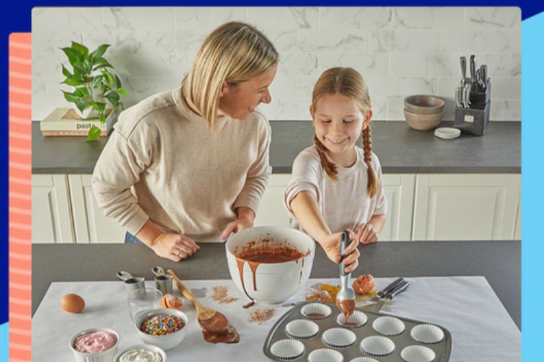 A woman and a girl enjoy baking together in a kitchen, with the girl smiling as she fills cupcake liners with chocolate batter from a bowl. Various baking ingredients are spread around, featured in the Reynolds Las Vegas Trip Sweepstakes