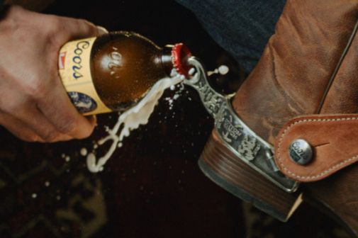 A hand pours a bottle of beer, causing froth to spill, while resting on the leather boot with a decorative spur, featured in the Coors Banquet x Dutton Ranch Earn Your Spurs Getaway