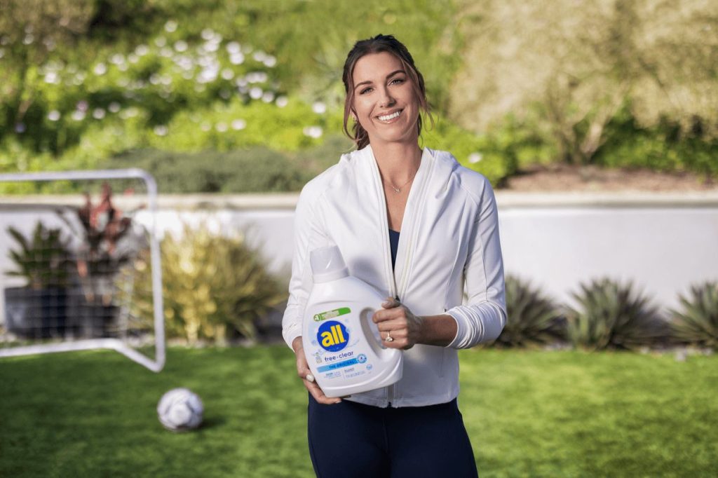 A smiling woman in a white zip-up jacket holds a laundry detergent bottle while standing on green grass, with a soccer goal and a ball visible in the background, featured in the all Most Valuable Parents Sweepstakes