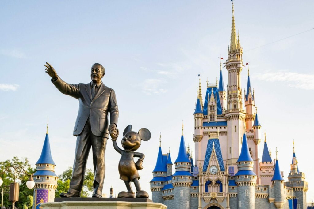 A bronze statue of Walt Disney and Mickey Mouse stands in front of a fairytale castle with blue turrets, under a clear sky, capturing a moment of imagination and wonder, featured in the Walt Disney World Coolest Summer Ever Sweepstakes