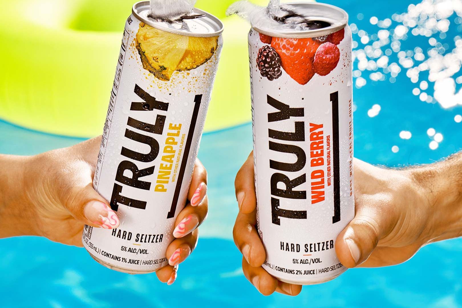 Two hands hold cans of pineapple and wild berry hard seltzers against a bright blue pool backdrop, capturing a moment of summer refreshment and enjoyment, featured in the Truly $10,000 Sweepstakes