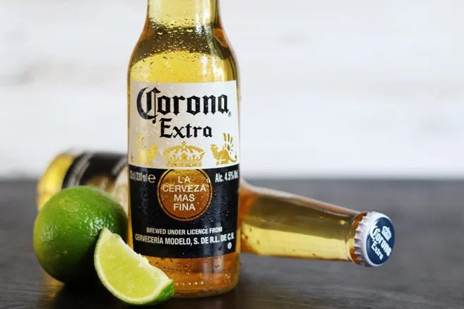 A chilled bottle of Corona Extra stands with a sliced lime on a dark surface, showcasing the refreshing beverage amid two additional bottles in the background, featured in the Corona Beach Getaway Sweepstakes