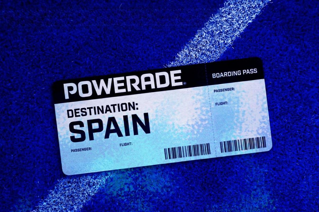 A boarding pass rests on a textured blue surface, indicating a destination of Spain, with the brand name Powerade prominently displayed at the top, featured in the POWERADE House of Legends Sweepstakes