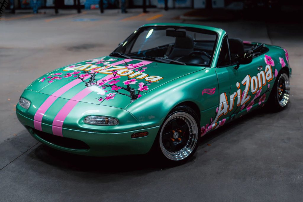 A vibrant green convertible car adorned with pink stripes and floral designs is parked in a garage, showcasing its unique artistic paint job and shiny wheels, featured in the AriZona Miata Sweepstakes