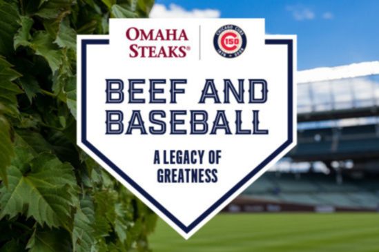 A vibrant baseball field is framed by lush greenery, showcasing a prominent sign featuring text about "Beef and Baseball" and celebrating a legacy of greatness, featured in the Omaha Steaks Cubs Trip Sweepstakes
