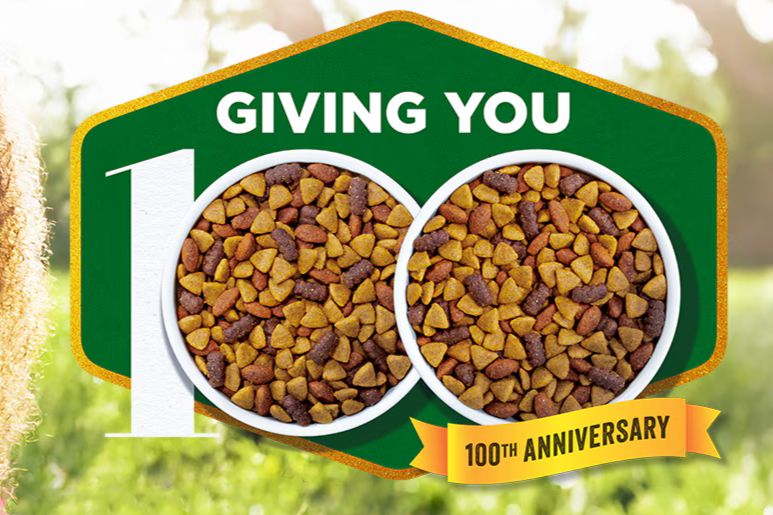 Two bowls of pet food filled with different colored kibble are centered in a vibrant outdoor setting, celebrating a 100th anniversary milestone with festive graphics, featured in the Purina $100,000 Sweepstakes