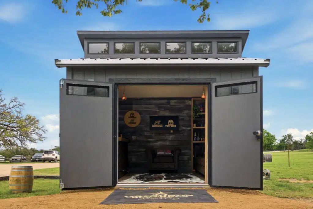A cozy, modern shed with open double doors showcases a rustic interior featuring wooden walls and furnishings, set against a clear blue sky and green landscape, featured in the Garrison Brothers Tuff Shed Sweepstakes