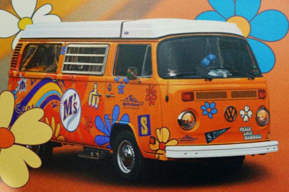 A bright orange Volkswagen van decorated with colorful flowers and playful graphics sits against a vibrant background, capturing a retro, cheerful aesthetic, featured in the Mariners 50 Seasons Van Sweepstakes
