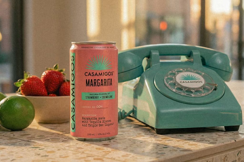 A vibrant pink can of margarita sits next to fresh strawberries and lime on a speckled countertop, alongside a retro turquoise telephone, illuminated by warm sunlight, featured in the Casamigos Phone Bill Sweepstakes