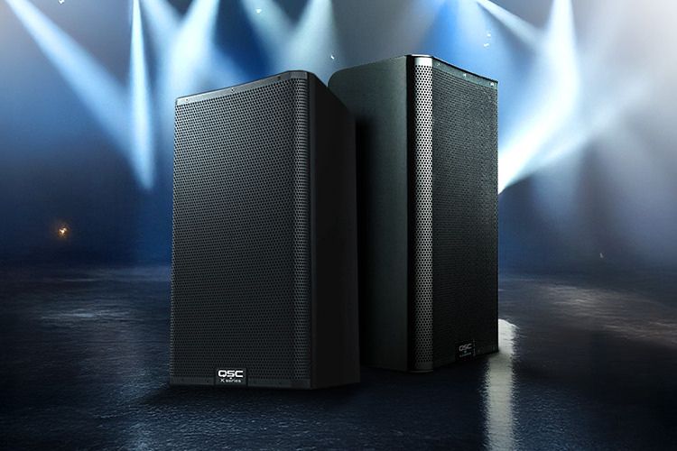 Two sleek black speakers stand prominently on a stage, illuminated by dramatic spotlighting, ready to deliver sound for a performance, featured in the Live Sound Month QSC Daily Giveaway