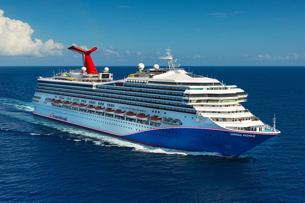 A large cruise ship sails through clear blue waters, showcasing its vibrant red and white design against a backdrop of fluffy clouds and open sky, featured in the Carnival Spring Sweepstakes