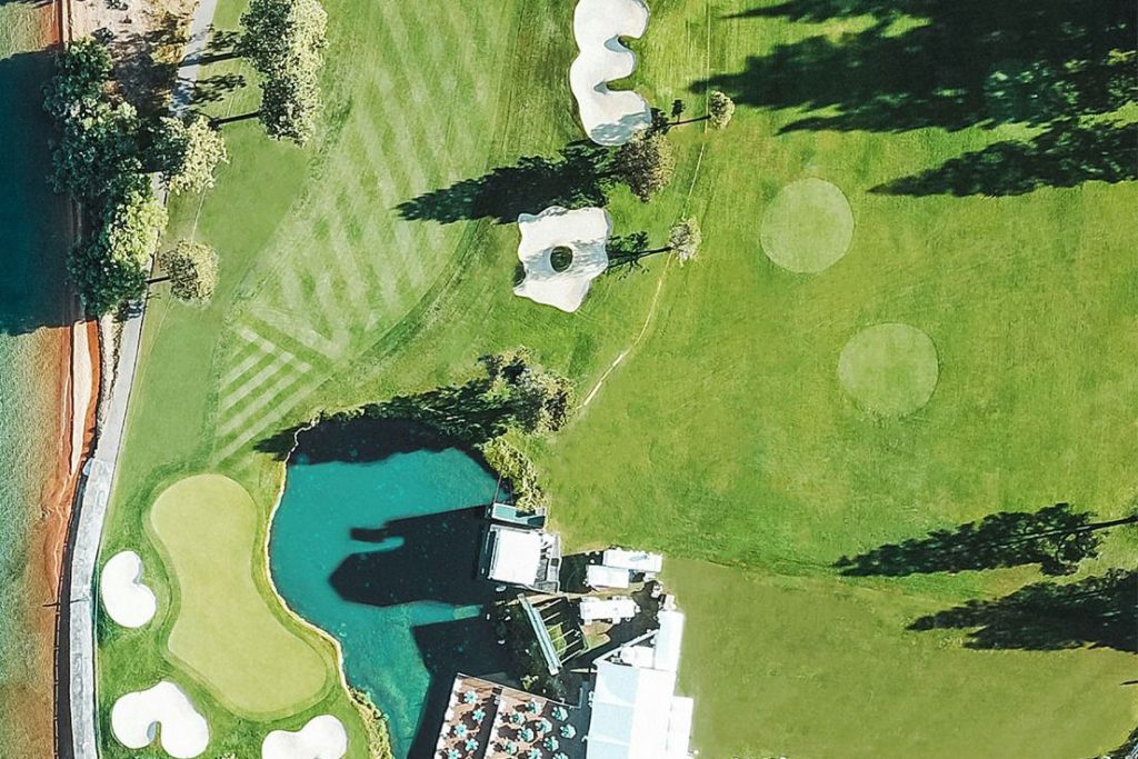 Aerial view of a picturesque golf course featuring manicured greens, bunkers, and a serene waterbody, surrounded by trees and a modern building with outdoor seating, featured in the Fujikura American Century Championship Sweepstakes
