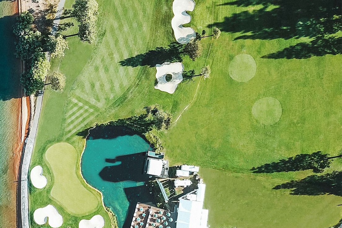 Aerial view of a picturesque golf course featuring manicured greens, bunkers, and a serene waterbody, surrounded by trees and a modern building with outdoor seating, featured in the Fujikura American Century Championship Sweepstakes