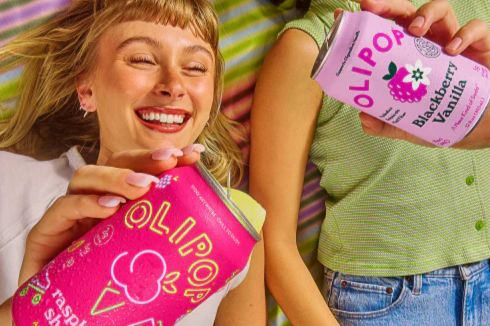 Two smiling young women hold colorful cans of sparkling beverages while lying on a striped blanket, enjoying a fun and refreshing moment together, featured in the Olipop Berry Sipport Sweepstakes