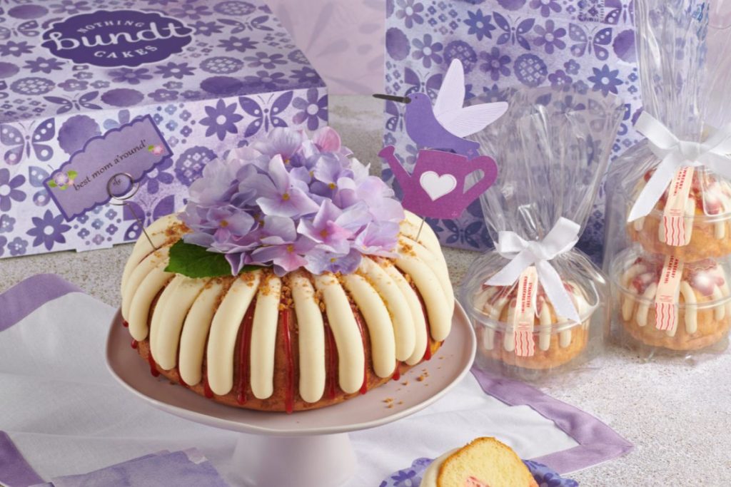A beautifully decorated bundt cake topped with vibrant purple flowers sits on a plate, surrounded by packaged mini cakes and floral-themed gift boxes, featured in the Nothing Bundt Cakes $4,000 Sweepstakes