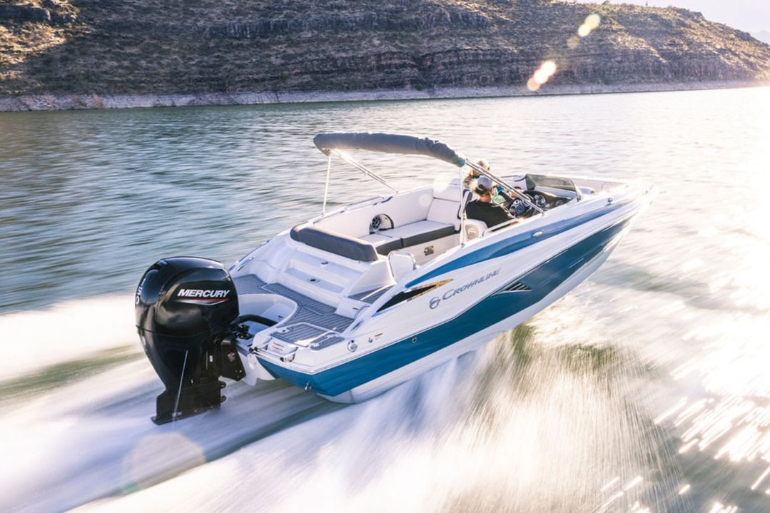 A speedboat glides across a shimmering lake, leaving a wake as a person steers confidently from the driver's seat. The rocky shoreline provides a scenic backdrop, featured in the Mercury Revive My Rig Giveaway