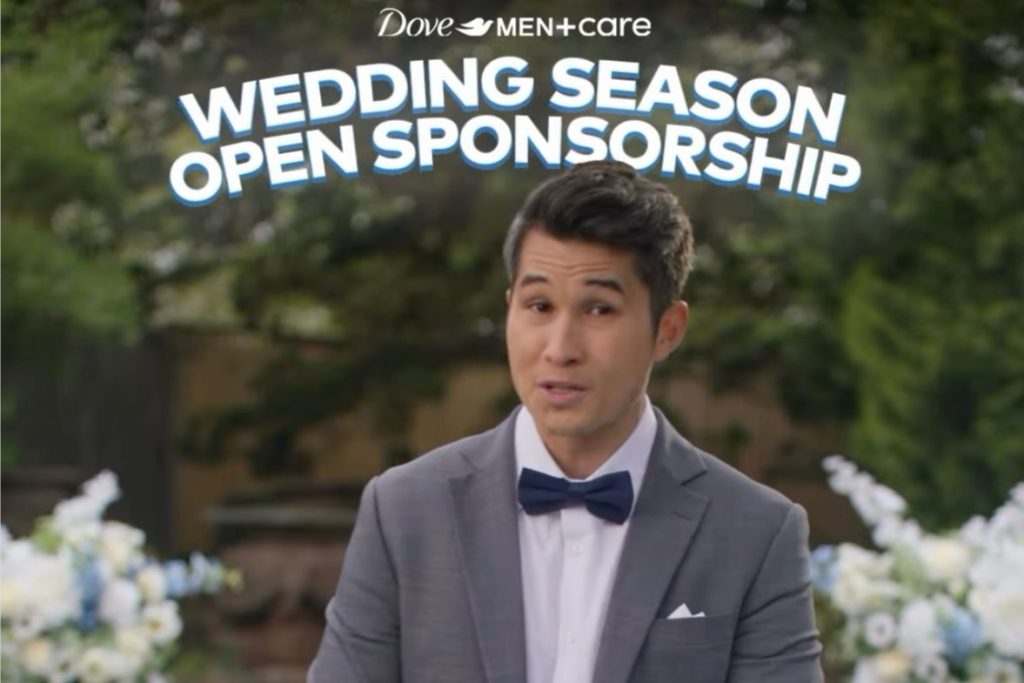 A well-dressed man in a gray suit and bow tie smiles as he speaks, with a backdrop of greenery and floral arrangements, suggesting a wedding theme, featured in the Dove Men+Care $50,000 Sweepstakes