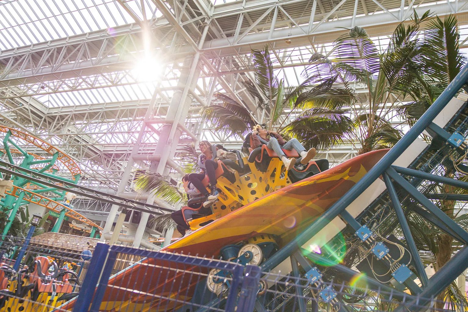 Thrill-seekers enjoy a roller coaster ride inside a vibrant indoor amusement park, surrounded by palm trees and bright sunlight filtering through the glass roof, featured in the Bloomington Minnesota $1,000 Mall of America Gift Card Sweepstakes