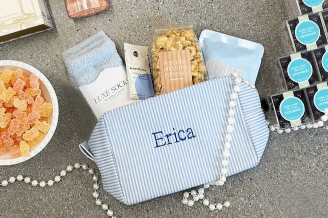 A cozy gift arrangement features a blue striped pouch with the name “Erica,” filled with luxurious socks, gourmet popcorn, skincare products, and colorful gummy candies on a textured surface, featured in the Sugarfina Bachelorette Gift Bag Sweepstakes