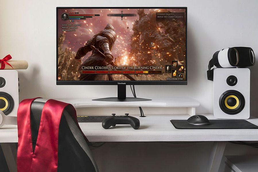 A gaming setup features a monitor displaying a battle scene with a character fighting the Cinder Colossus, accompanied by a gaming controller and speakers on a white desk, featured in the ViewSonic Graduation Sweepstakes