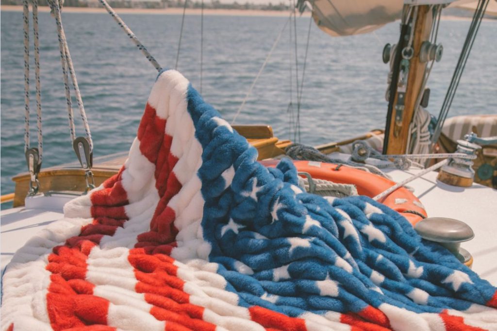 A cozy red, white, and blue blanket drapes over a boat deck, with calm water and a distant shoreline visible, creating a serene maritime atmosphere, featured in the Minky Couture Flag Blanket Sweepstakes