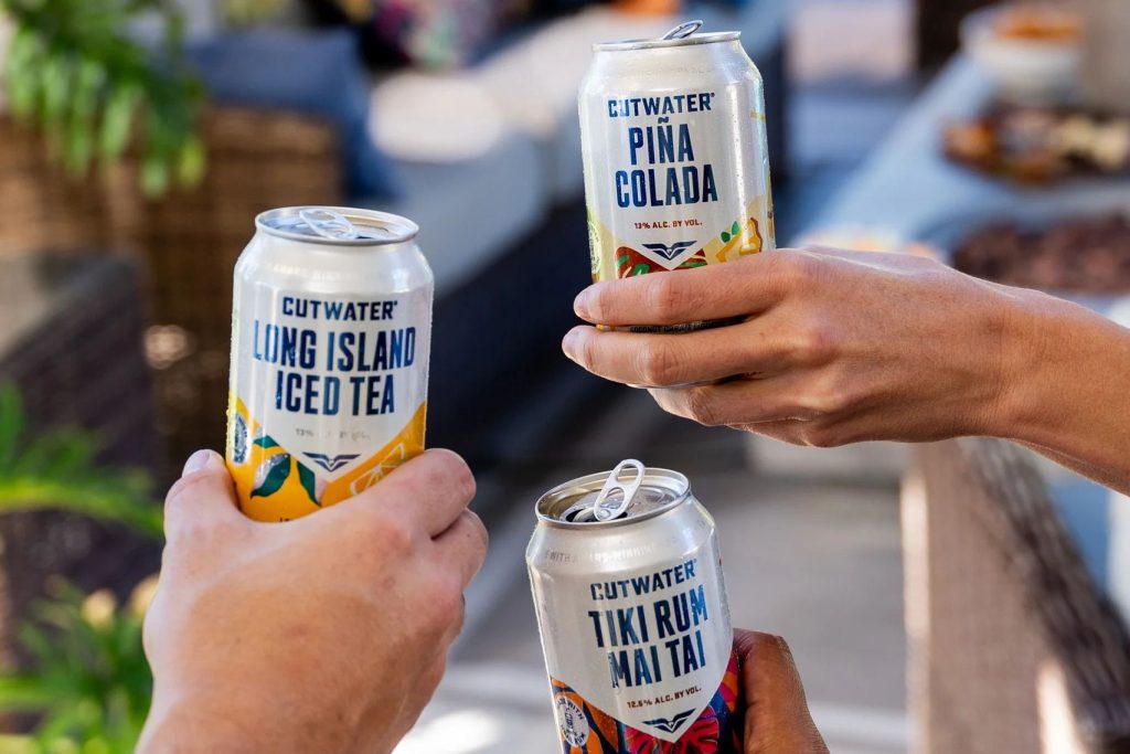 Three hands hold cans of mixed drinks, showcasing Long Island Iced Tea, Piña Colada, and Tiki Rum Mai Tai, in a lively outdoor setting with blurred background elements, featured in the Cutwater Tiki Getaway Sweepstakes