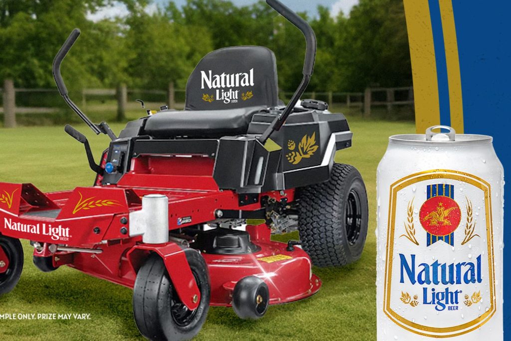 A red riding lawnmower stands on a grassy field, featuring a black seat adorned with the words "Natural Light." A cold can of beer sits prominently beside it, featured in the Natural Light Zero Turn Mower Sweepstakes