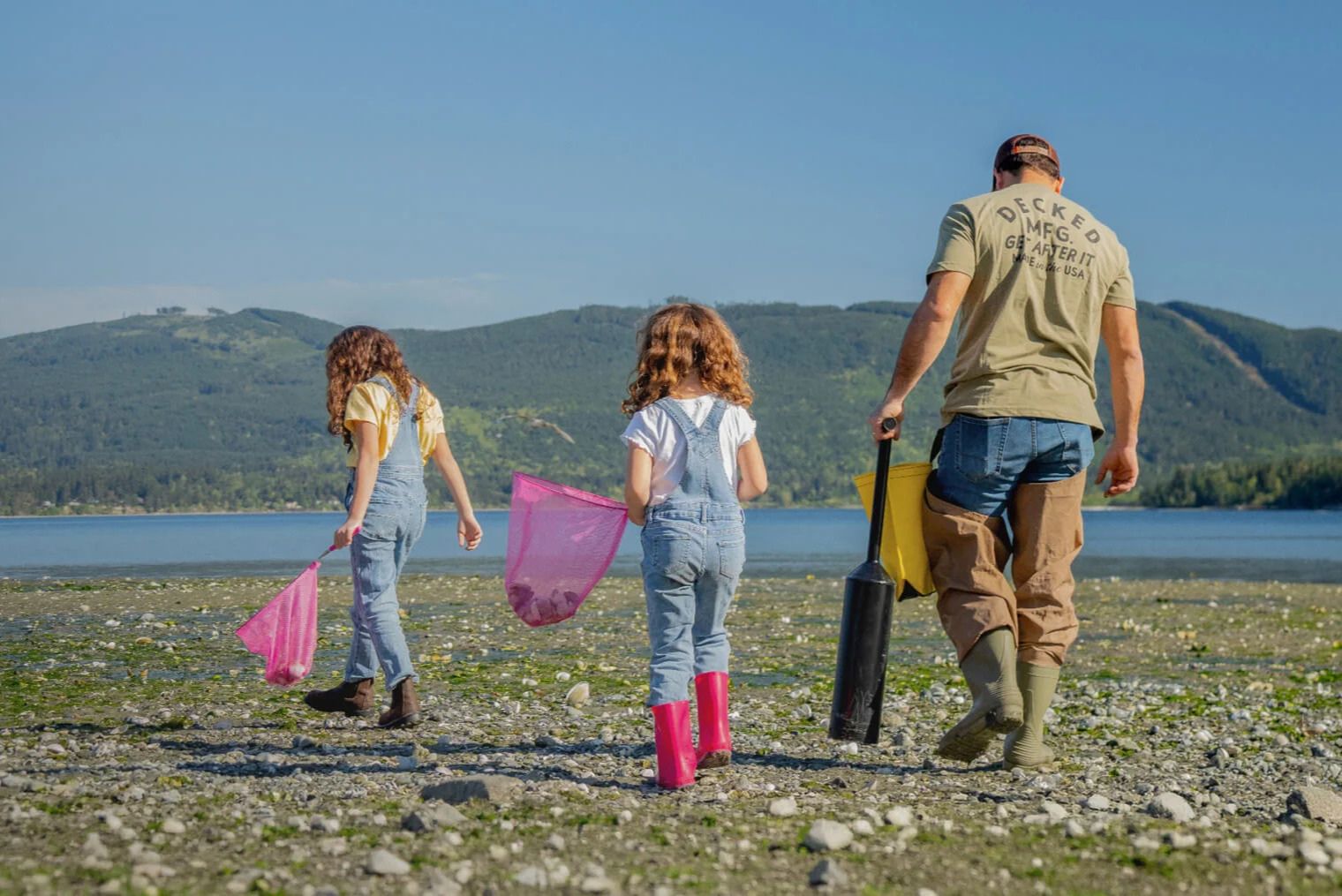 A man and two girls explore a rocky shoreline, each holding colorful nets and walking towards the water. They are surrounded by green hills under a clear blue sky, featured in the DECKED Dad Time Off Sweepstakes