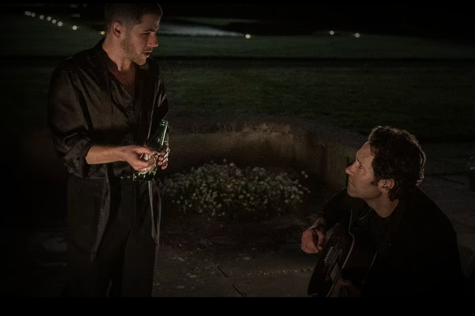 Two men engage in conversation at night; one holds a green bottle while the other plays guitar, surrounded by a softly lit outdoor setting and greenery, featured in the How To Write A Song Sweepstakes