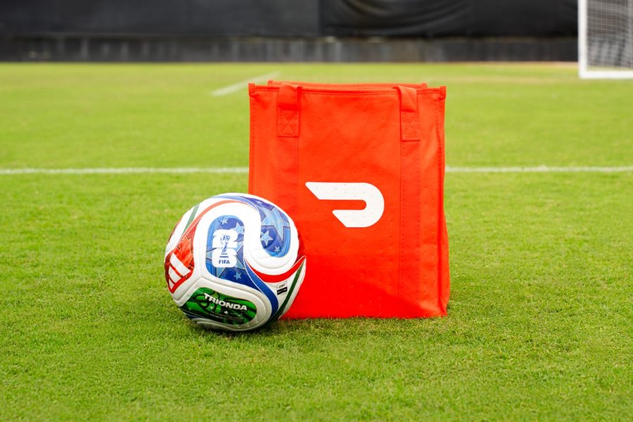A soccer ball rests on the green field next to a bright red bag, emphasizing a sporty scene that hints at upcoming athletic activity, featured in the DoorDash FIFA World Cup Final Sweepstakes