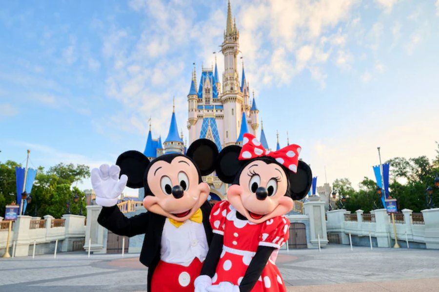 Mickey and Minnie Mouse pose joyfully together in front of a majestic castle, showcasing their signature outfits and charming smiles against a clear blue sky, featured in the Disney+ Cool Kids Summer Sweepstakes