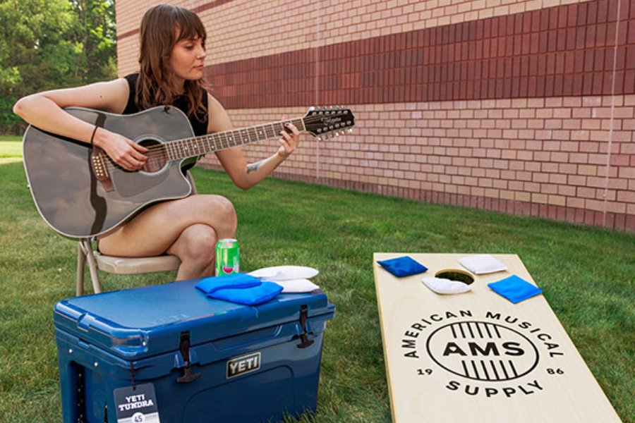 A young woman plays acoustic guitar while seated on a chair outdoors, with a cooler beside her and a cornhole game set up in front. Green grass and a brick wall provide the backdrop, featured in the American Musical Supply Sweepstakes