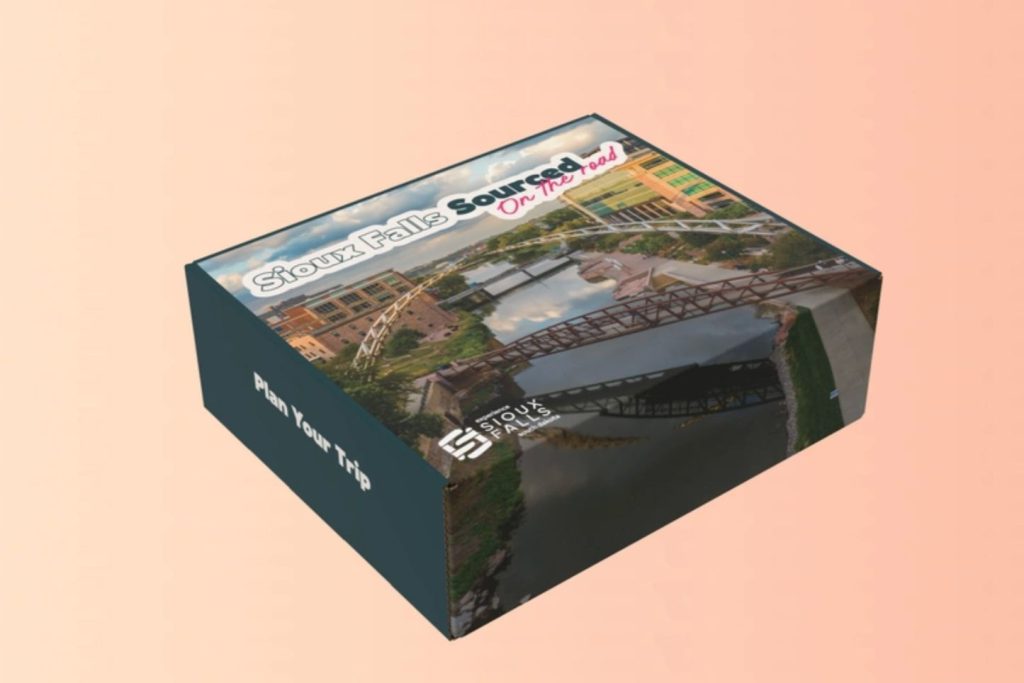 A colorful box featuring a scenic view of Sioux Falls, showcasing bridges and water. The text invites viewers to plan their trip, highlighting the area's attractions, featured in the Experience Sioux Falls Sourced Box Sweepstakes
